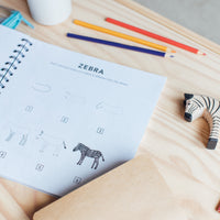 Open spiral bound book of "All the Animals: How to Draw Books" by Alli Koch showing step-by-step zebra illustration on a wooden desk with colored pencils, a toy zebra, and sketching supplies in a cozy playroom setting