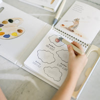 Open book with coloring pages and a hand holding a pen on a light surface.