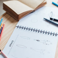 A little notebook sitting top of of an open spiral bound book of "All the Things: How to Draw Book for Kids (Spiral)" showing step-by-step instructions on how to draw a rocket and a planet. Pens and colored pencils around it.