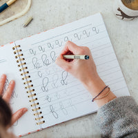 Person practicing calligraphy in a notebook with a pen and coffee mug on a table.
