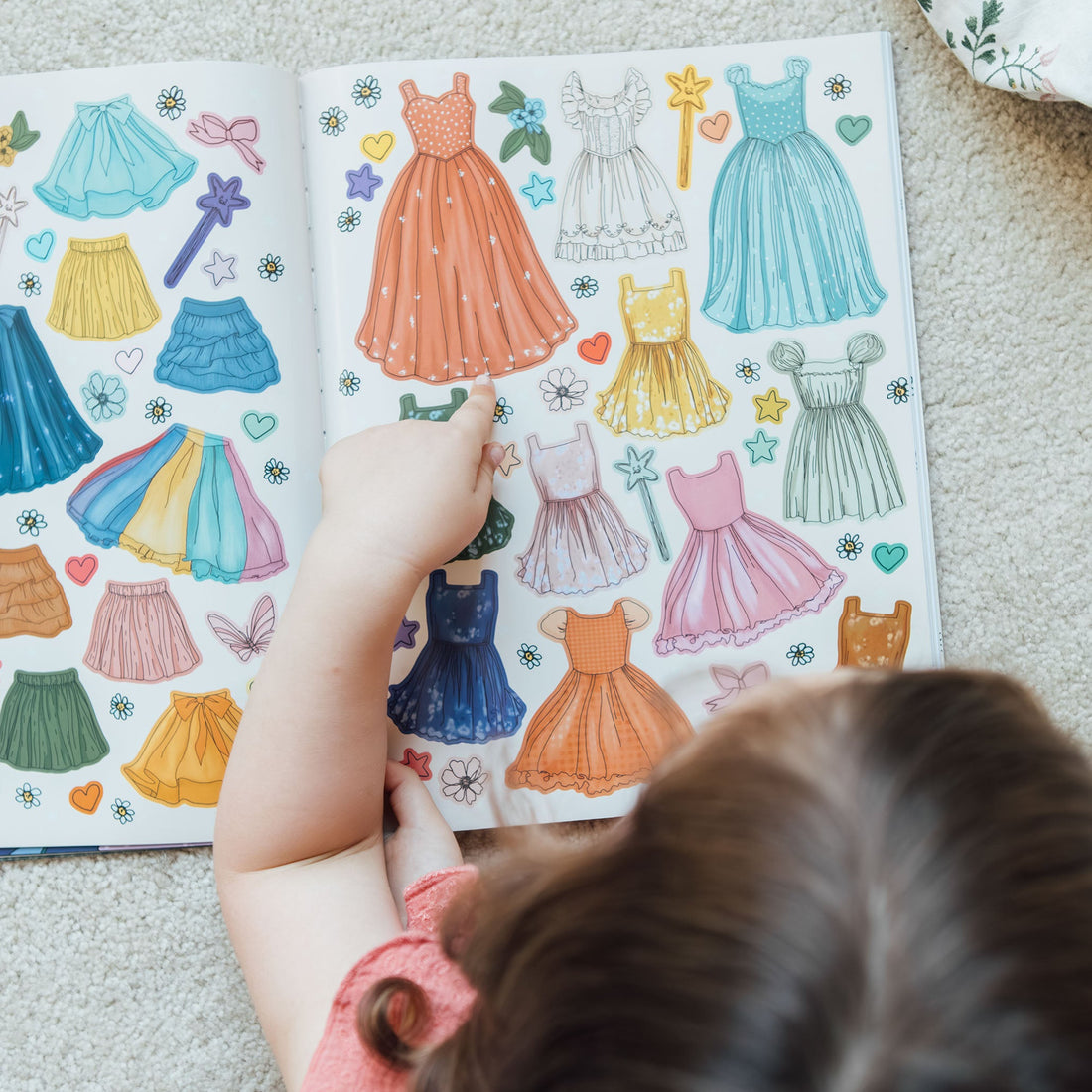 Child looking at a sticker book with colorful dress illustrations on a carpeted floor.