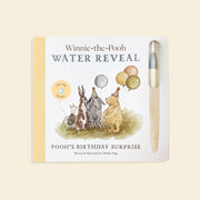 Winnie-the-Pooh Water Reveal book with a wooden stick and a small round disc, shown on a light cream background. The book cover features Winnie-the-Pooh characters with balloons and the title 'Winnie-the-Pooh WATER REVEAL'.