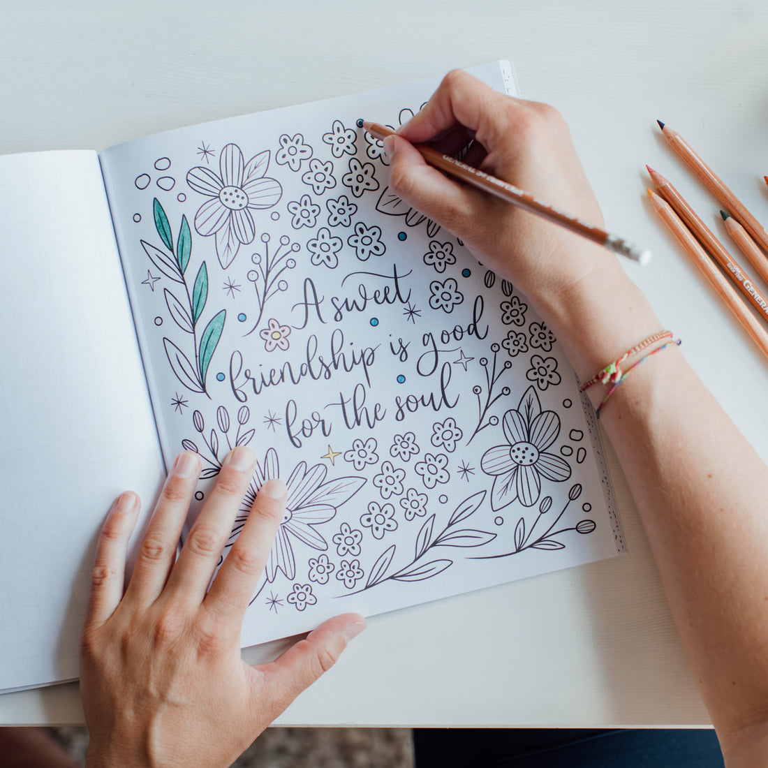 person coloring in an open book of 'Coloring Faith'. the page shows floral line art and a text 'A Sweet friendship is good for the soul'.