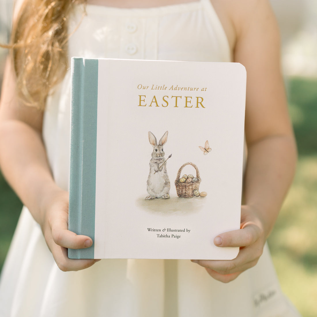 Young girl holding a book titled 'Our Little Adventure of Easter' with an illustration of a rabbit and basket on the cover.