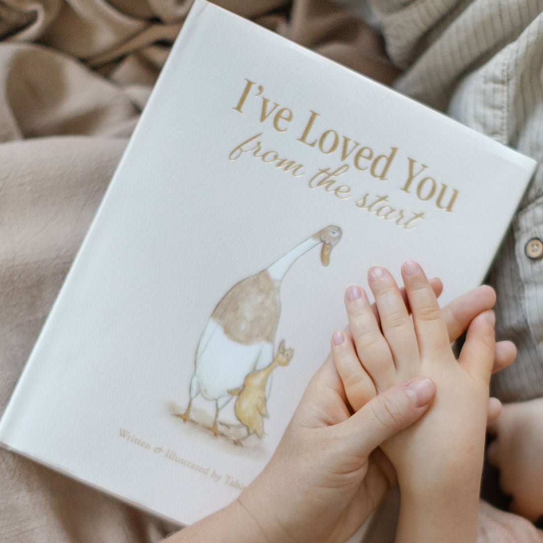 A children's book titled "I've Loved You from the start" held in hands over beige fabric, with a soft illustration of a mother goose and goose on the cover.