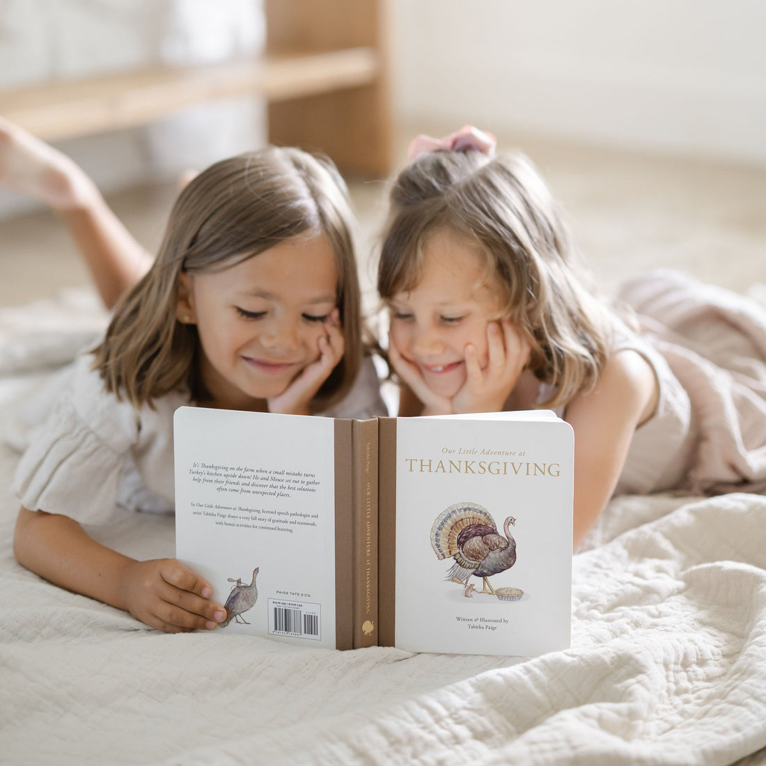 Two young girls lying on a quilted bed, smiling while looking at a personalized children's book titled 'The Little Thanksgiving' with a turkey illustration on the back cover.
