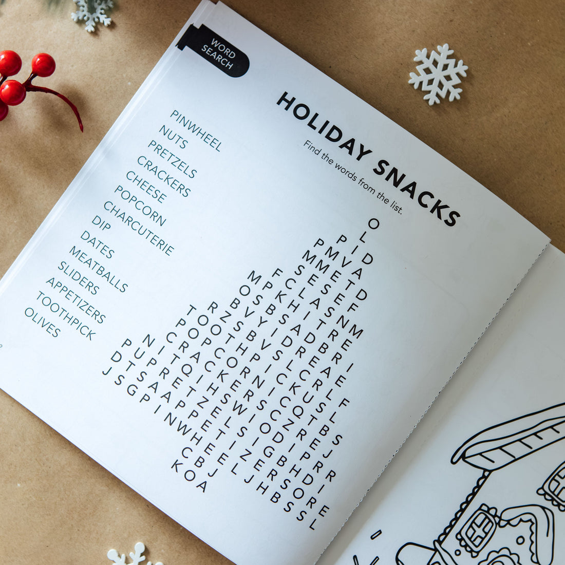An open printed holiday-themed word search page titled 'HOLIDAY SNACKS' laid on a kraft paper surface with Christmas decorations around it, including evergreen branches, red berries, white snowflakes, and ornaments.