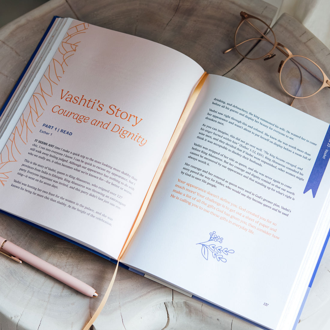 An open book titled 'Vashti's Story: Courage and Dignity' lies on a light stone surface, styled with a pink pen, gold-rimmed eyeglasses, and a thin orange ribbon bookmark. The book pages are white with orange and blue text and illustrations.