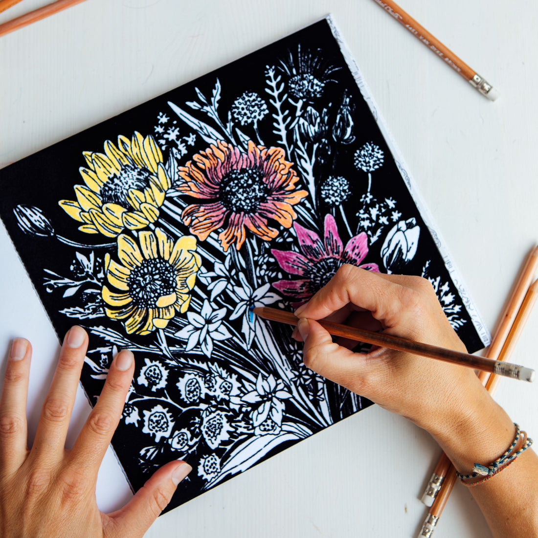 A hand holds a pencil over a square canvas featuring a floral linocut or print with yellow, orange, and pink flowers on a black background, placed on a white surface with colored pencils around it.
