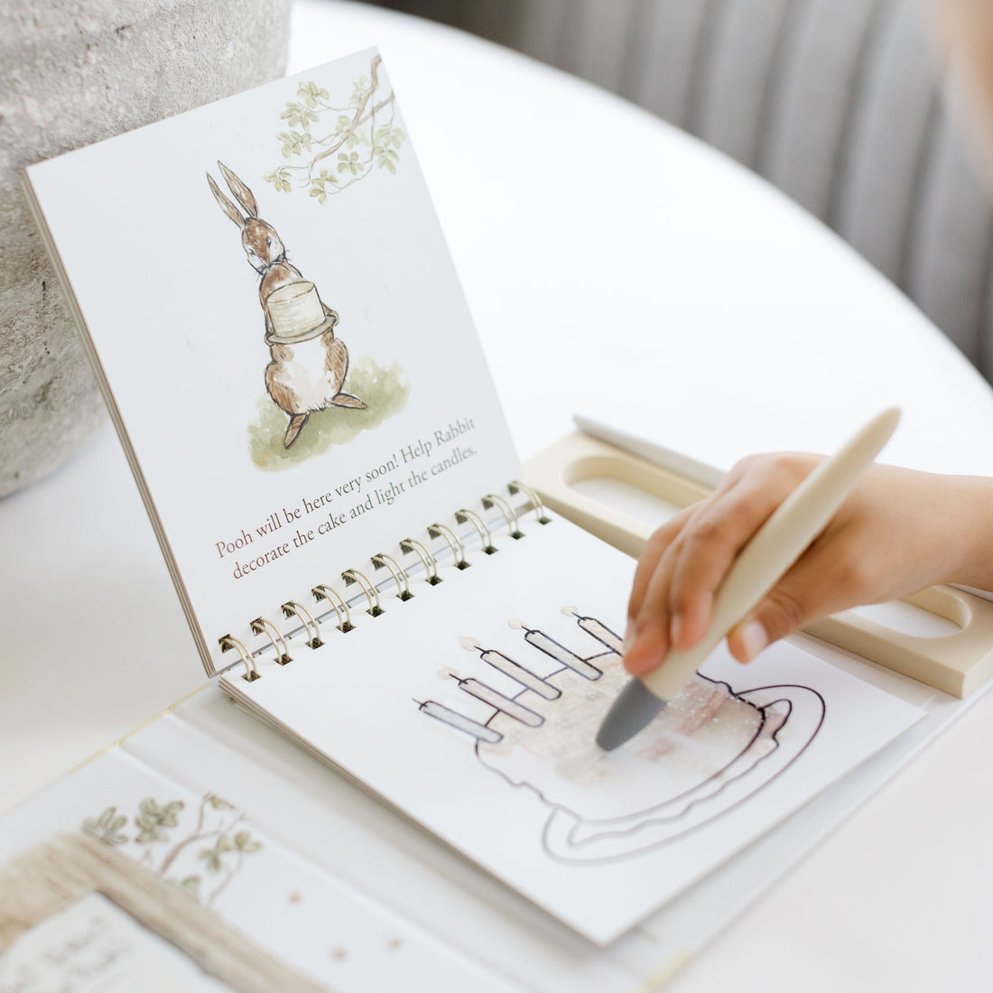 A child's hand holding a wooden paintbrush and coloring a page in a spiral-bound children's activity book. The book features a watercolor rabbit illustration on the cover and is placed on a white table next to a textured gray vase.