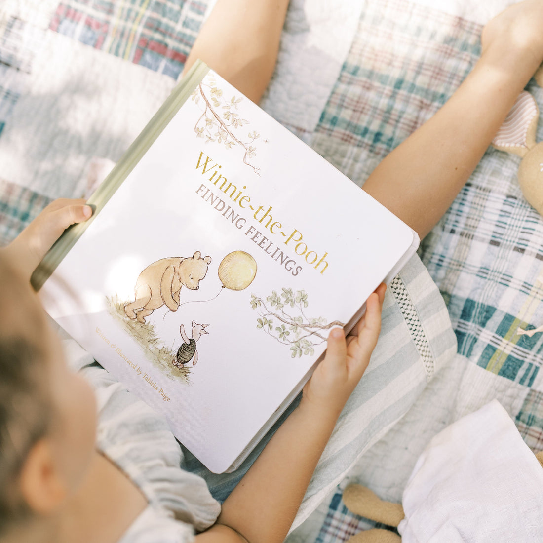 A child holds a large illustrated book titled "Winnie-the-Pooh Finding Feelings" while sitting on a plaid blanket. The book cover shows Winnie-the-Pooh and a rabbit with a yellow balloon, and the photo is taken in a soft, sunlit indoor setting.