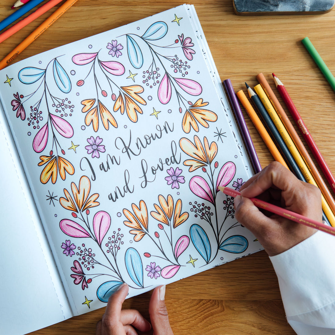 open book of 'Fearless & Faithful' shows a colored in coloring page of flowers with the text "I am know and loved". A person is coloring the page with a colored pencil. 