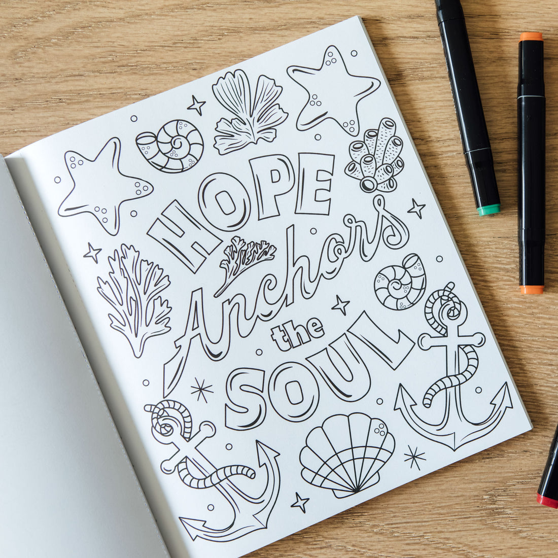 A black-and-white coloring page with nautical illustrations and the words “HOPE Anchors the SOUL,” placed on a wooden surface beside several markers and a small glass bowl.