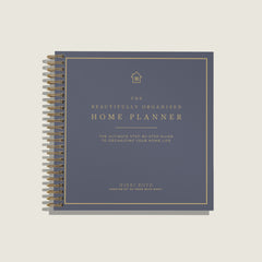 A spiral-bound home planner book titled 'The Beautifully Organized Home Planner' by Nikki Boyd, featuring a slate blue cover with gold text and border details, shown on a light beige background.