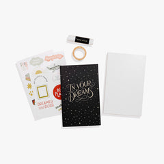 A vision board kit laid out on a white background, including a guided journal, a blank board, photos, stickers, hand-drawn patterns, decorative washi tape, and a non-toxic glue stick.